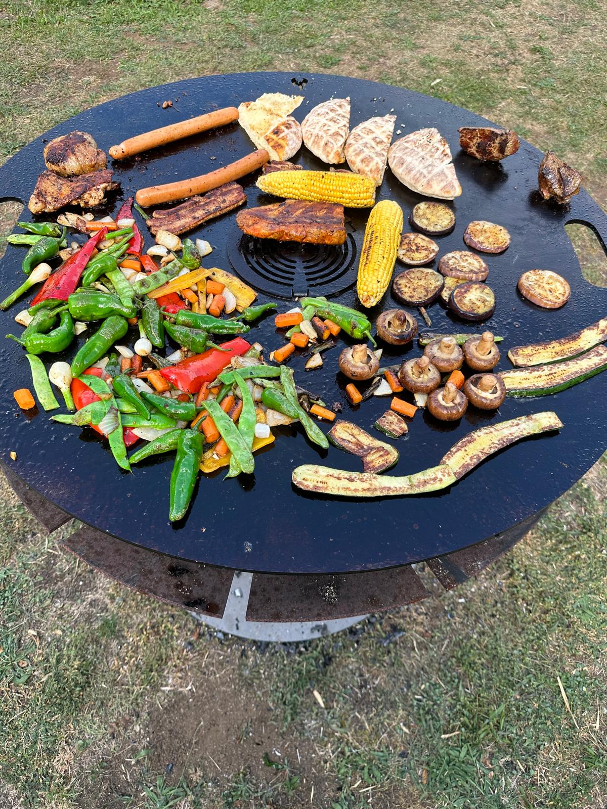 Feuerplatte Ø800 mm im Einsatz mit Grillgemüse, Fleisch und Outdoor-Cooking über offenem Feuer